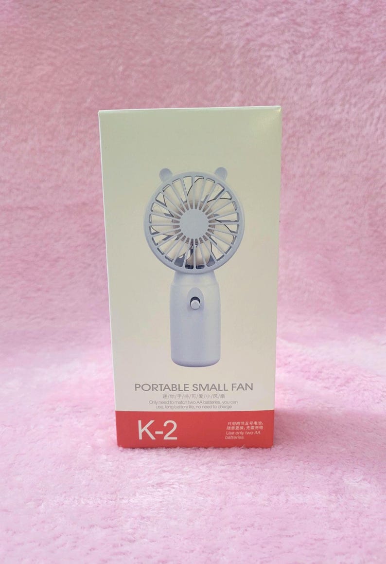 Op de afbeelding: Een witte draagbare kleine ventilator met een beren-vormige bovenkant, verpakt in een witte doos. De ventilator heeft een ronde kop met bladen en een handvat. De doos heeft de tekst "PORTABLE SMALL FAN" en "K-2".