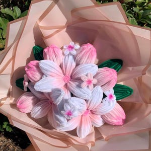 May include: A bouquet of handmade flowers in shades of pink and white, with green leaves, wrapped in light pink tissue paper. The flowers have a soft, textured appearance, and the arrangement is tied together with a ribbon.
