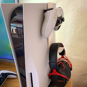May include: A white PlayStation 5 console with a black controller and red headphones hanging on the side. The console is on a wooden surface.