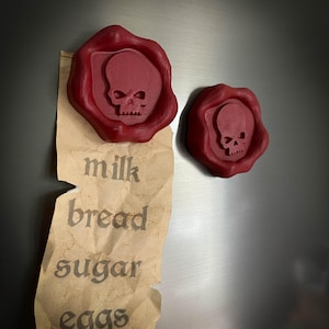 Op de afbeelding: Twee rode, schedelvormige zegelwasmagneten. Eén is bevestigd aan een bruin papieren boodschappenlijstje met de woorden "milk, bread, sugar, eggs". De andere magneet is rechts geplaatst. De magneten bevinden zich op een zilveren oppervlak.