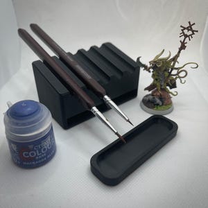 Könnte beinhalten: Ein schwarzer Plastik-Pinselhalter mit zwei braunen Pinseln, einer kleinen schwarzen Farbpalette und einer blauen Flasche Citadel Colour Macragge Blue Farbe.