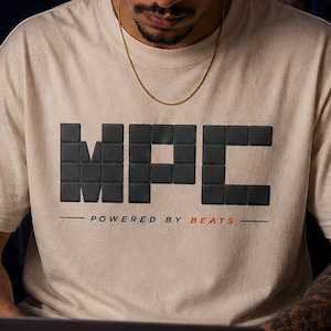 Może przedstawiać: Beżowa koszulka z napisem "MPC" w bloku liter, z napisem "POWERED BY BEATS" poniżej. Osoba ma na sobie czarną czapkę i złoty łańcuszek. Na biurku znajduje się laptop i czarny kubek.