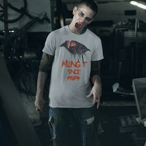 K&ouml;nnte beinhalten: Ein Mann tr&auml;gt ein wei&szlig;es T-Shirt mit einem Grafik von Freddy Krueger aus dem Film "Nightmare on Elm Street" und dem Text "Killing It Since 1984". Er tr&auml;gt zerrissene blaue Jeans und hat einen bedrohlichen Gesichtsausdruck.