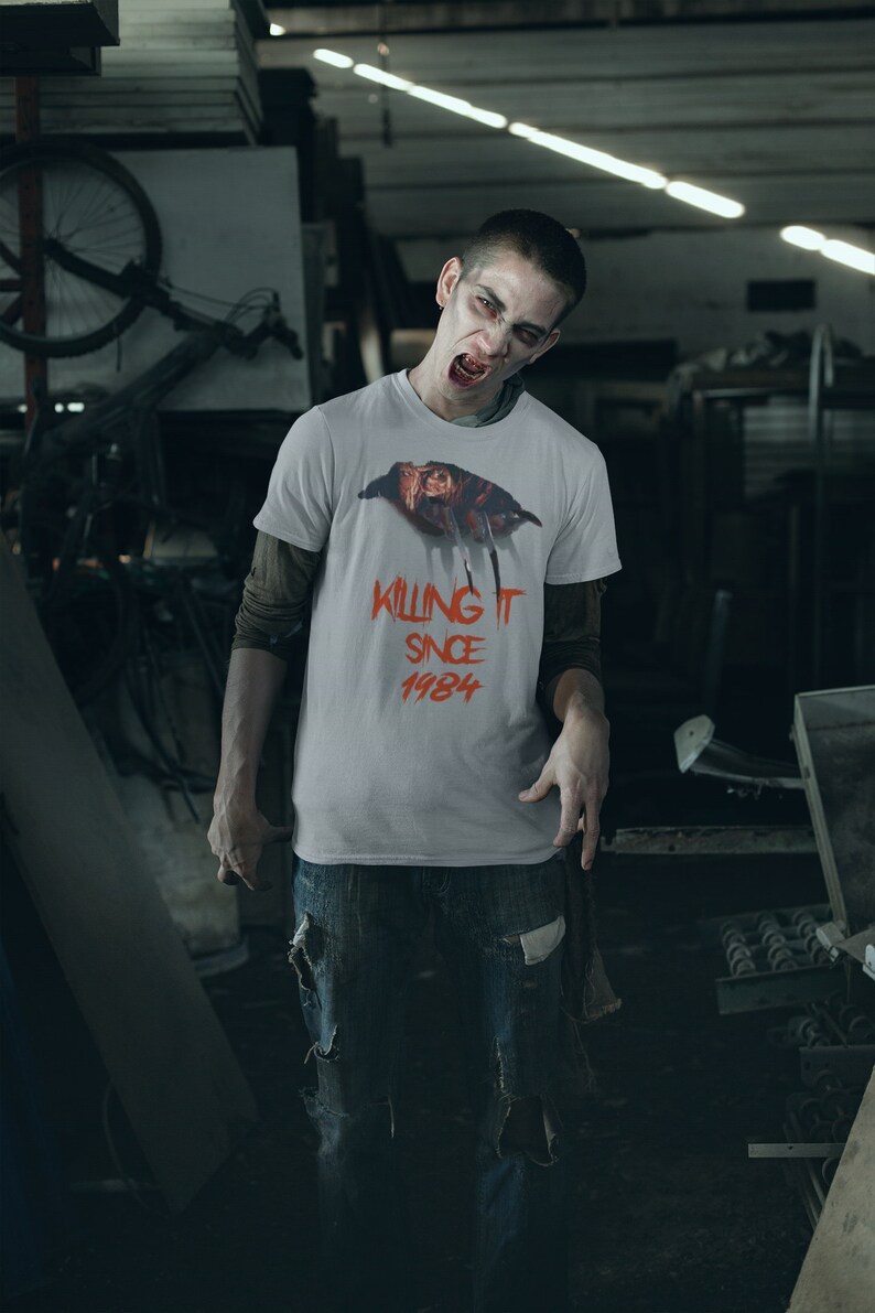 K&ouml;nnte beinhalten: Ein Mann tr&auml;gt ein wei&szlig;es T-Shirt mit einem Grafik von Freddy Krueger aus dem Film "Nightmare on Elm Street" und dem Text "Killing It Since 1984". Er tr&auml;gt zerrissene blaue Jeans und hat einen bedrohlichen Gesichtsausdruck.