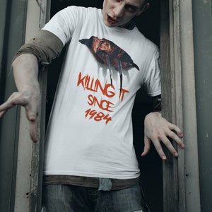 K&ouml;nnte beinhalten: Ein wei&szlig;es T-Shirt mit einer roten Grafik eines gruseligen Gesichts und dem Text "Killing It Since 1984".