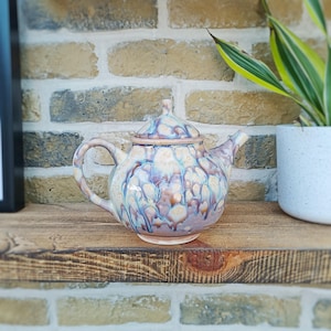 May include: A ceramic teapot with a light brown, white and purple marbled glaze. The teapot has a round body and a spout on the side. The teapot is sitting on a wooden shelf.