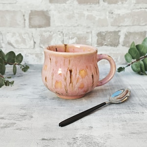May include: A pink ceramic mug with a textured, speckled glaze and a brown handle. The mug is sitting on a white wooden surface.