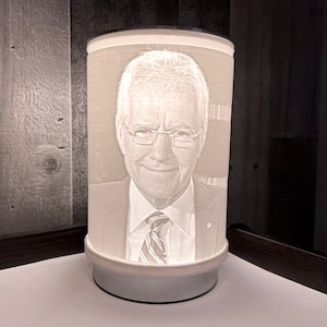 Pode incluir: Lâmpada cilíndrica branca com um retrato de um homem de terno e gravata. A lâmpada tem uma base e uma borda superior. O retrato é gravado na superfície da lâmpada e a lâmpada é iluminada por dentro.