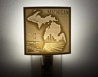 Op de afbeelding: Een nachtlampje met een houten uitsnijding van de staat Michigan met het woord "MICHIGAN" bovenaan. De uitsnijding bevat een brug, een skyline en golven.