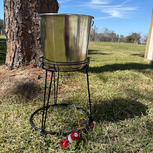 May include: A silver metal bucket with handles sits on a black metal stand with a black hose and red gas regulators. The stand is on a green grassy lawn.