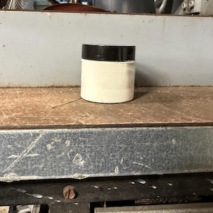 Könnte beinhalten: Ein zylinderförmiger Behälter mit schwarzem Deckel und weißem Körper. Der Behälter steht auf einer Holzoberfläche. Der Hintergrund ist eine Metallstruktur.