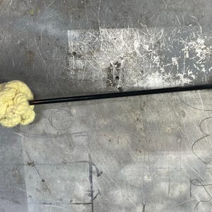 May include: A black metal rod with a yellow, fluffy, round object attached to one end. The rod has a metal ring at the other end.