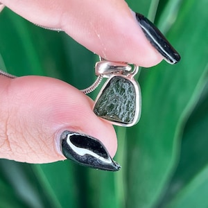 May include: A pendant featuring a raw, dark olive-green moldavite stone with a textured, irregular surface set in a polished silver-toned metal frame. The pendant is attached to a thin, flexible silver-toned chain necklace.