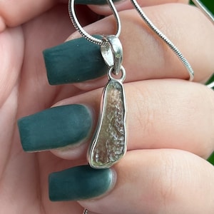 May include: A silver pendant necklace featuring a raw, textured, light brown stone set in a smooth silver bezel. The pendant hangs from a thin, flexible silver snake chain, showcasing a natural, irregular shape with a rough, pitted surface texture.