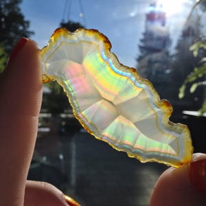 May include: A translucent, irregularly shaped agate slice with a rainbow iridescence. The edges are a warm brown, transitioning to a white and clear center. The stone is held up to the light, showcasing its natural patterns.