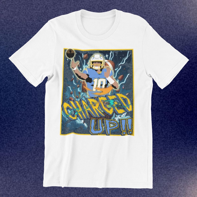 May include: White t-shirt featuring a graphic of a football player in a blue and yellow uniform, throwing a football. The design includes lightning bolts and the words "CHARGED UP!!" in yellow and blue.