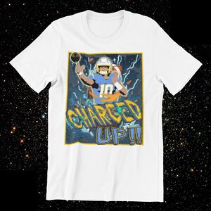 May include: White t-shirt featuring a graphic of a football player in a blue and yellow uniform throwing a football. The text "CHARGED UP!!" is displayed in a stylized font below the player.