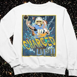 May include: White sweatshirt featuring a graphic of a football player in a blue and yellow uniform, with the text "CHARGED UP!!" in yellow and blue. The design includes lightning bolts and a football.