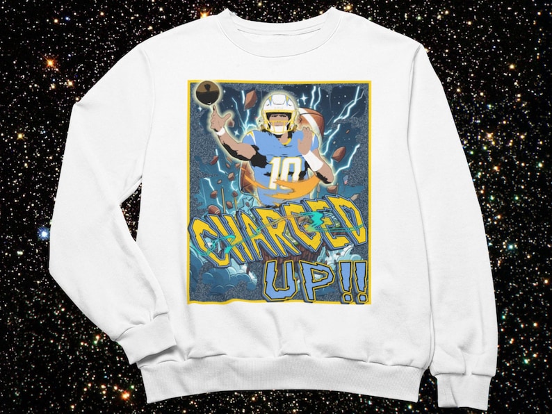 May include: White sweatshirt featuring a graphic of a football player in a blue and yellow uniform, with the text "CHARGED UP!!" in yellow and blue. The design includes lightning bolts and a football.