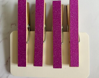 May include: Four wooden clothes pegs with a pink glitter finish. The clothes pegs are arranged in a row on a white surface.