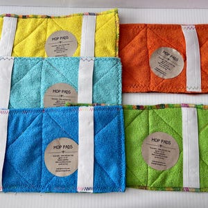 May include: A selection of colourful mop pads in yellow, blue, turquoise, orange, and green. Each pad features a white strap and a circular label that reads "Mop Pads". The pads are made of a textured fabric.