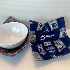 May include: Two fabric bowl cozies featuring Penn State University branding with blue, white, and grey colours, including paw prints and text logos. One cosy contains a plain white ceramic bowl with a ribbed exterior, while the other is empty.