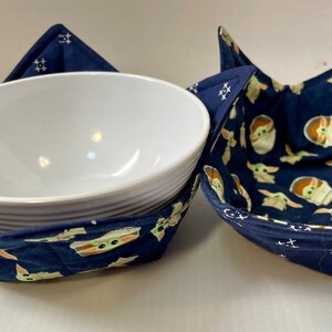 May include: Two blue fabric bowl cosies with a pattern of green baby Yoda characters. The cosies are folded and a white bowl is inside one of the cosies.