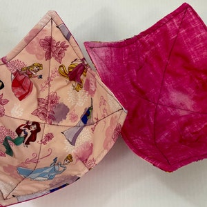 May include: Two fabric bowl cosies with square, quilted designs. One cosy features a light pink background with a repeating pattern of various animated princess characters and floral motifs. The second cosy is a solid, textured dark pink fabric.