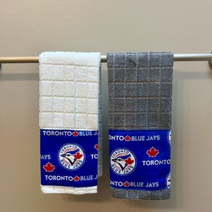 May include: Two hanging hand towels. One is white, the other is gray. Both have a blue band with the text "TORONTO BLUE JAYS" and a logo featuring a bird and a maple leaf. The towels are hanging on a silver bar.