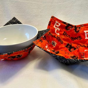 May include: Two bowl cozies with a white bowl in one. The cozies are orange with white and black text and designs. The text says "BC Lions" and "CFL". The other cozy is black with white designs.