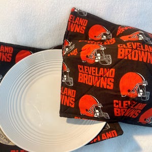 May include: A fabric bowl cosy featuring a black background with a repeating pattern of orange football helmets and the text Cleveland Browns in orange. A white, circular, ribbed ceramic bowl is positioned inside the quilted fabric holder.