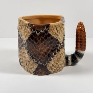 May include: A ceramic mug featuring a textured rattlesnake skin pattern in shades of tan and dark brown. The handle is shaped like a segmented rattlesnake tail with brown, black, and white bands, extending outward from the side of the mug.