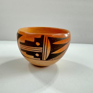 May include: A handcrafted, round ceramic bowl with a light brown base and a vibrant orange rim. The bowl features geometric black and orange designs around the exterior. The interior is a warm orange color.