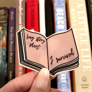 May include: A sticker in the shape of an open book with the text "long story short" on the left page and "I survived" on the right. The book has a pink interior and blue and yellow page edges. The sticker is held in front of a bookshelf.