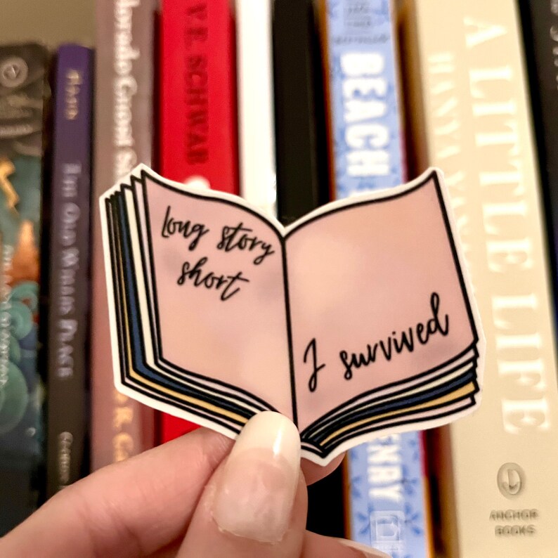 May include: A sticker in the shape of an open book with the text "long story short" on the left page and "I survived" on the right. The book has a pink interior and blue and yellow page edges. The sticker is held in front of a bookshelf.