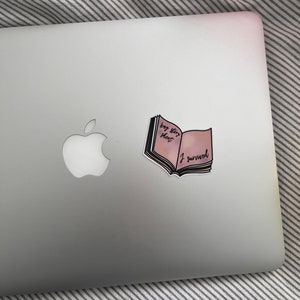 May include: A sticker in the shape of an open book with the text "big story short I survived" on a silver laptop. The book has pink and yellow pages with black text. The laptop is on a striped surface.