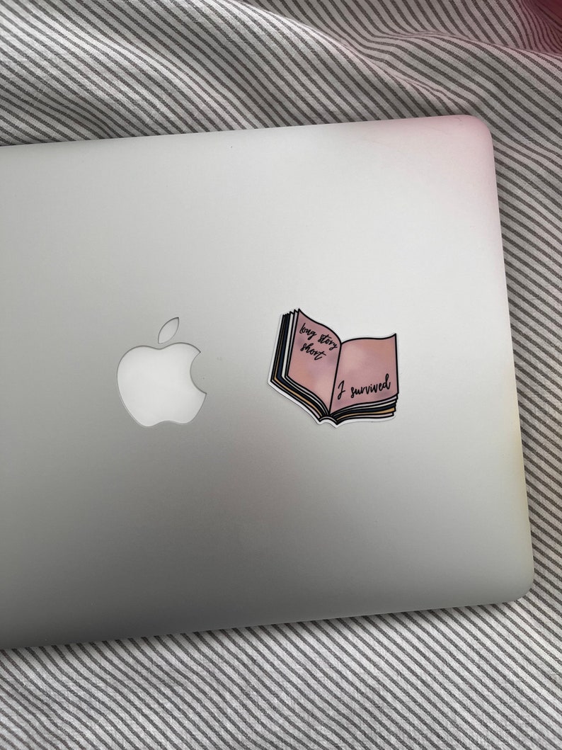 May include: A sticker in the shape of an open book with the text "big story short I survived" on a silver laptop. The book has pink and yellow pages with black text. The laptop is on a striped surface.