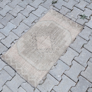 Puede incluir: Una alfombra rectangular en tonos beige y gris claro, con un patrón geométrico central en forma de diamante y un diseño de borde sutil. La alfombra tiene una textura de pelo corto y bordes deshilachados en los lados cortos, aspecto desgastado.