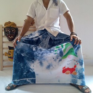 May include: A man wearing a white shirt and a blue and white patterned skirt with a gnome design. He is sitting on a wooden chair with a wooden mask behind him.
