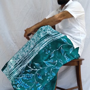 May include: A man wearing a white shirt sits on a wooden chair. He is wearing a long, teal blue sarong with a white and blue batik pattern. The sarong has a floral design with white stems and blue leaves.