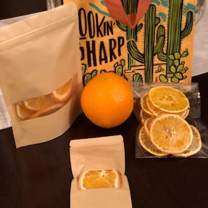 Puede incluir: Tres bolsas de papel marrón con rodajas de naranja en su interior. Las bolsas están sobre una superficie de madera marrón oscuro. Una bolsa tiene una sola rodaja de naranja dentro, otra tiene varias rodajas de naranja y la tercera tiene una naranja entera junto a ella. Todas las bolsas están selladas con una cremallera.