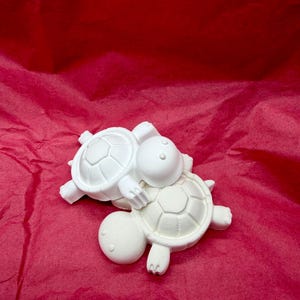 May include: Two white turtle figurines stacked on top of each other. Each turtle features a smooth, rounded head with two small circular eyes and a detailed shell with a hexagonal pattern on the back. The figures are solid white in color.