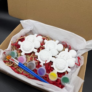 May include: A craft kit in a cardboard box containing five white turtle-shaped plaster figures, two blue-handled paintbrushes, and six small circular containers filled with various colors of paint, all nestled on red and brown paper shred.