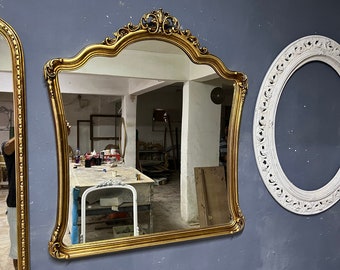 May include: A gold-framed mirror with an ornate, curved top is mounted on a grey wall. The mirror reflects a workshop interior. A second mirror with a similar gold frame is visible on the left. A white oval frame is on the right.