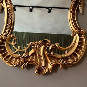 May include: A gold-colored ornate mirror with a decorative, intricate frame. The frame has a scalloped design with a raised, swirling pattern.