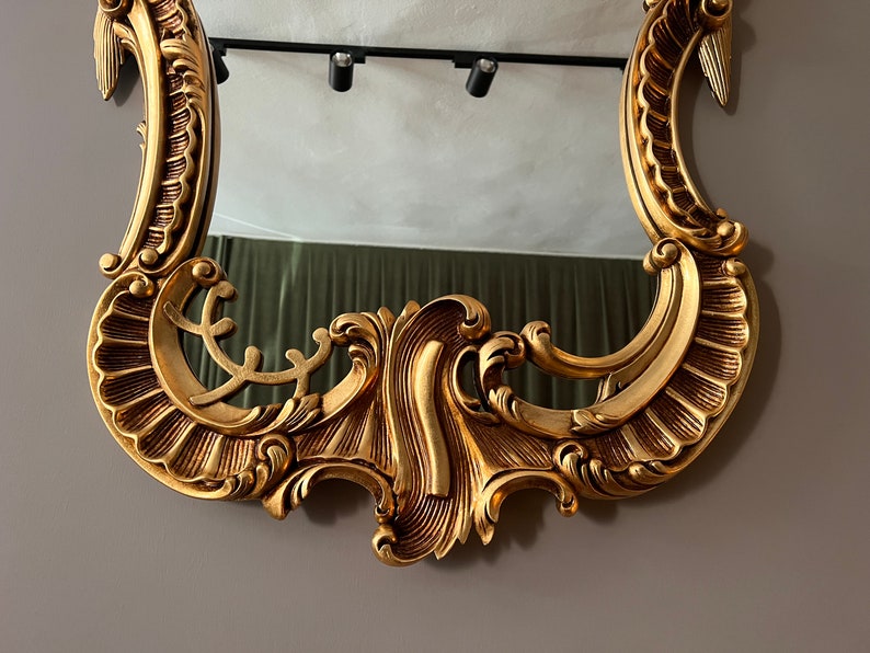 May include: A gold-colored ornate mirror with a decorative, intricate frame. The frame has a scalloped design with a raised, swirling pattern.