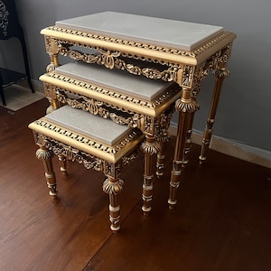 May include: A set of three gold-painted nesting tables with white marble tops. The tables are intricately carved with floral designs and have a classic, ornate style.