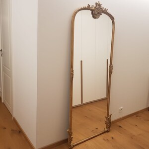 Op de afbeelding: Een hoge, sierlijke gouden spiegel met een decoratief bovenstuk en een gefacetteerde rand. De spiegel staat in een kamer met witte muren en houten vloeren.