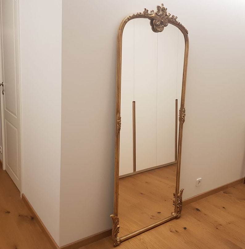 Op de afbeelding: Een hoge, sierlijke gouden spiegel met een decoratief bovenstuk en een gefacetteerde rand. De spiegel staat in een kamer met witte muren en houten vloeren.