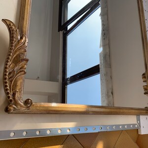 Op de afbeelding: Een sierlijke gouden spiegel met een decoratief, gerold frame. De spiegel reflecteert een raam met uitzicht op de lucht.